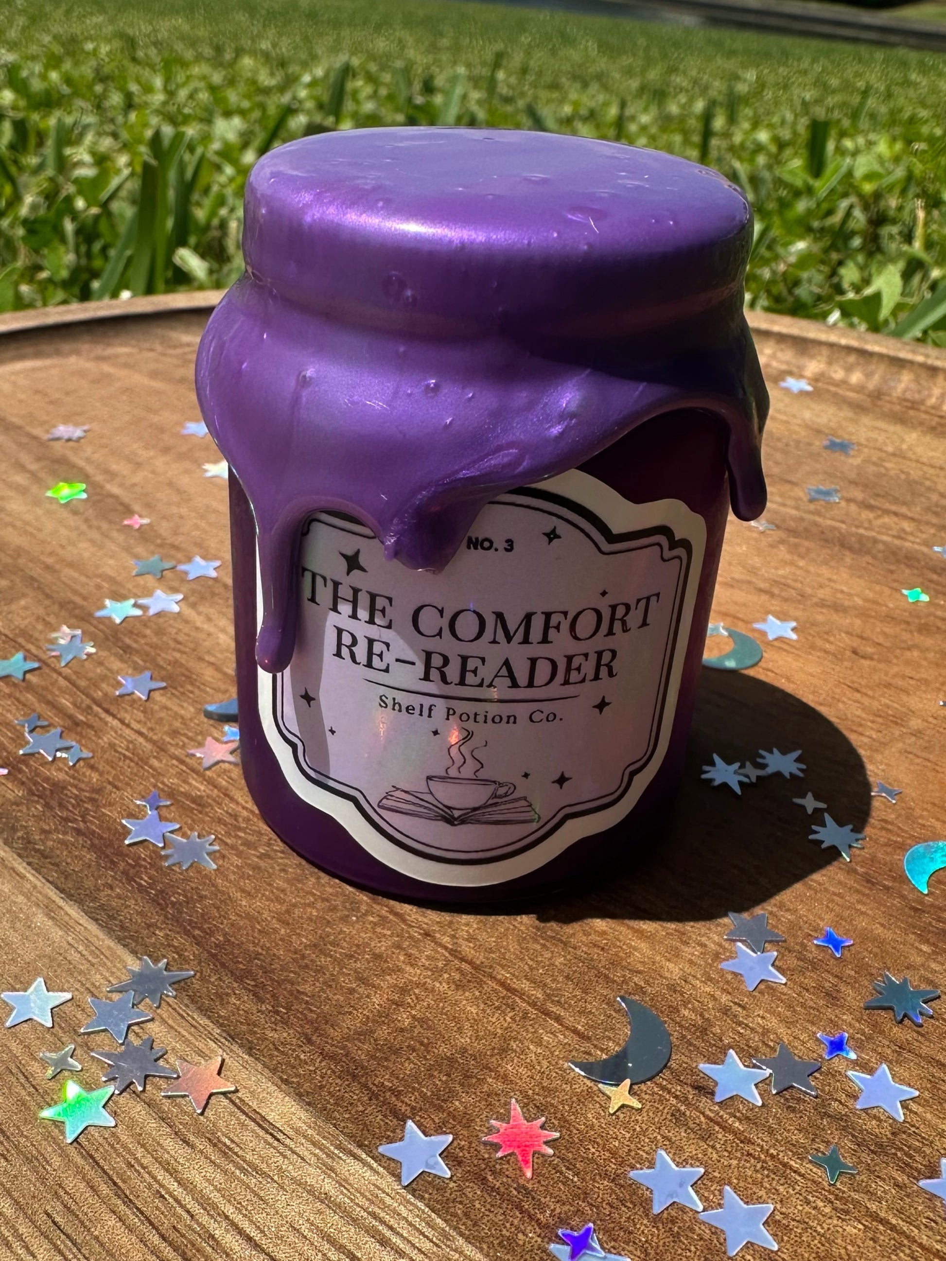 Purple bottle labeled 'The Comfort Re-Reader' on a wooden surface with star-shaped confetti.
